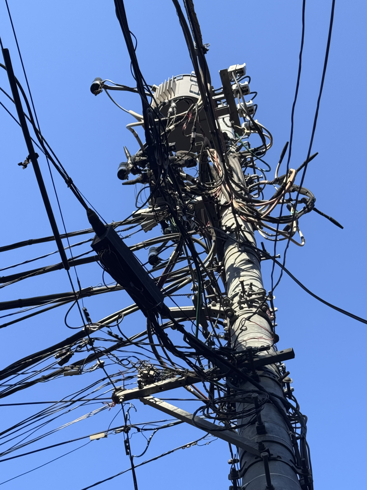 Dettaglio dal basso di un palo a Tokyo, con apparecchiature e un fitto groviglio di cavi contro il cielo azzurro.